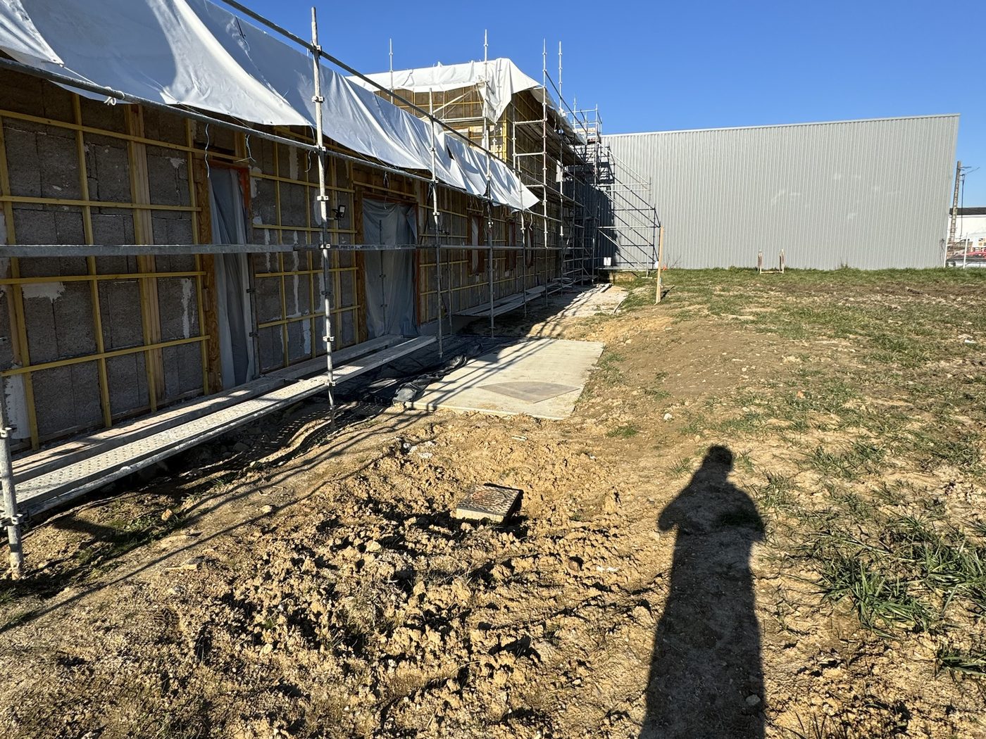 Terrain en terre nue devant une façade en chantier, avec échafaudage et bâche