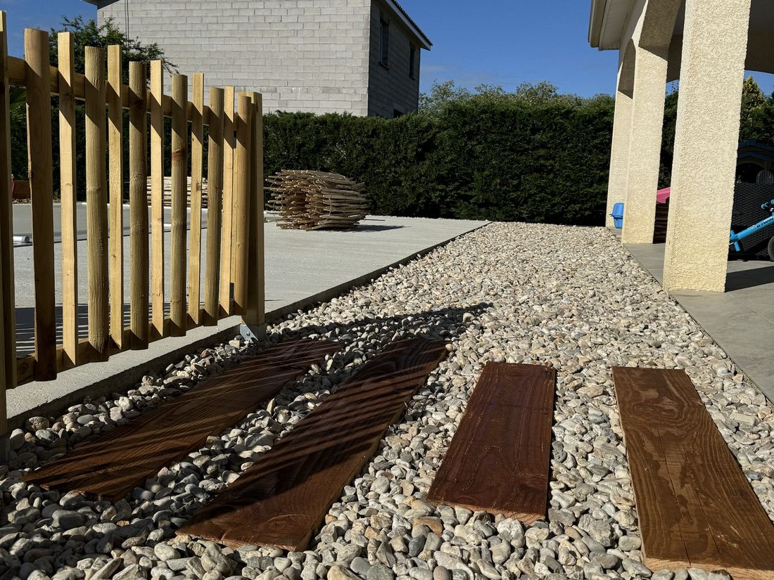 Clôture en bois à lattes verticales, allée de galets et dalles en bois, devant une maison à arcades