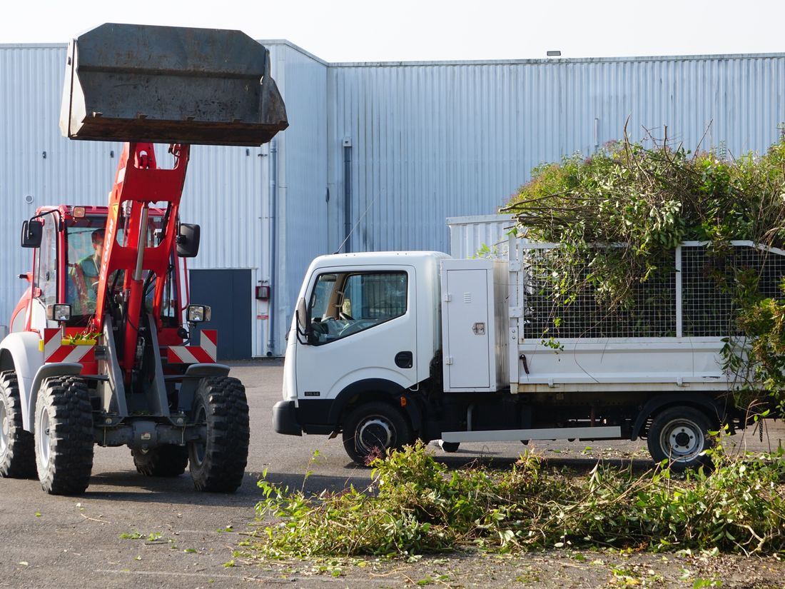 Chargeuse et camion benne grillagé chargés de déchets verts devant un bâtiment industriel