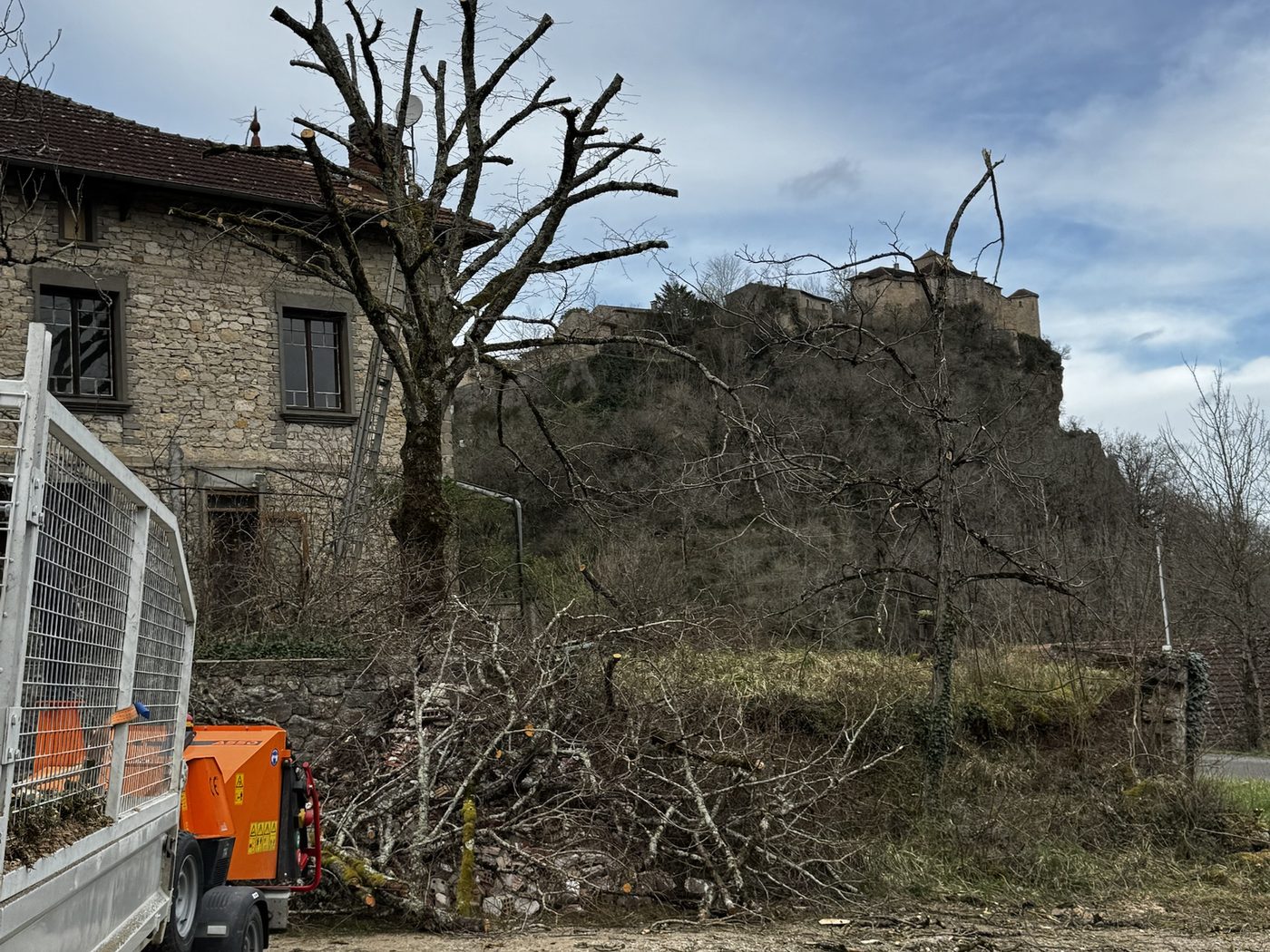 Arbre élagué devant une maison en pierre, avec broyeur de branches et camion benne