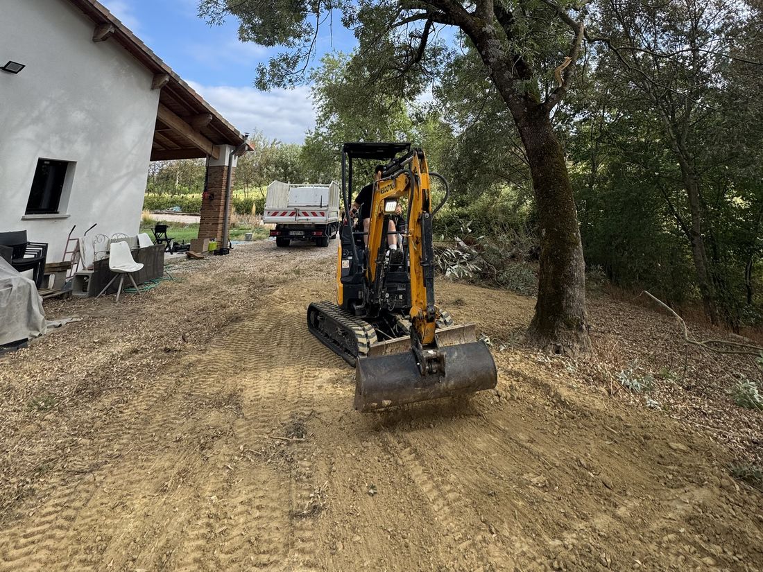 Mini-pelle sur chenilles au pied d'un grand arbre, devant une maison, avec un camion benne en arrière-plan